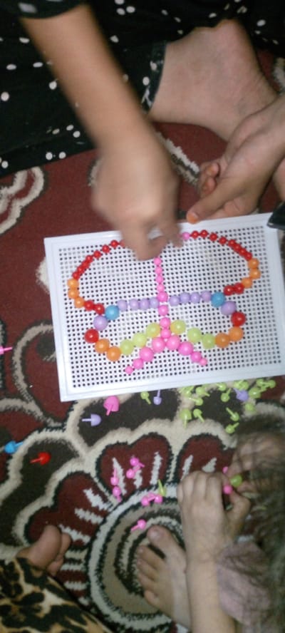 In Marhamat, Uzbekistan, a child is engaged in a bead art activity on a white pegboard. The pegboard, filled with colorful beads, is positioned on a patterned rug. The child's hands are visible, actively placing beads to form a butterfly shape. Scattered around the pegboard are additional loose beads and peg-like accessories, suggesting an ongoing creative process. The scene depicts a common childhood pastime, highlighting a moment of focused play within a domestic setting.