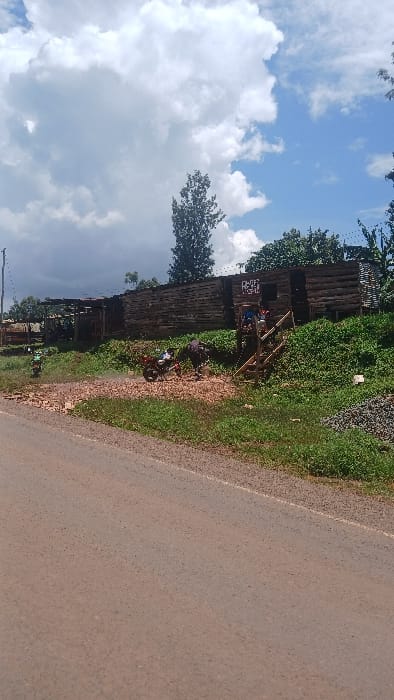 The scene depicts an outdoor view in Boochi Tendere ward, Kenya, under a partially cloudy sky. A reddish-brown dirt road occupies the foreground, running horizontally across the lower portion of the image. An overgrown green embankment rises sharply from the road's edge.

On the embankment, to the right, a prominent long wooden structure, likely a building or large kiosk, is constructed from rough-hewn logs or timber, elevated on a foundation. An entrance to this structure is accessible via a set of wooden steps. A partially visible sign, displaying letters "RVAF" among others, is affixed to the front of this building. Three individuals are visible near this entrance: one person stands at the top of the steps, and two others are seated or standing on the steps or just inside the doorway. Their specific clothing details are not clearly discernible.

To the left of the wooden steps, a red and black motorcycle is parked on a patch of light-brown wood chips or debris, which is spread across the ground. A bag or backpack is secured to the rear of this motorcycle. Further to the left, a green motorcycle is partially visible near additional, less defined wooden structures. A pile of dark, coarse material, possibly gravel or charcoal, is visible on the far right of the embankment.

The background features several trees, including a tall, slender tree centrally located. Lush green vegetation covers much of the embankment and surrounding areas. A utility pole with wires is visible on the far left. The overall environment appears rural or semi-urban.