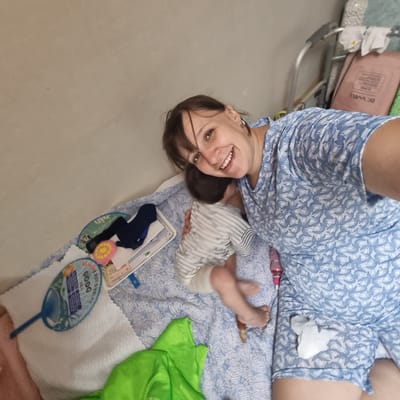 A woman and a baby are lying on a bed. The woman is wearing a blue and white floral pattern pajama set and is smiling at the camera. The baby is wearing a striped onesie and is lying on its stomach, facing away from the camera. There are various objects on the bed, including a blue fan with Korean text, a pink and yellow scrubber, a white tray with some items, and a green piece of fabric. The background appears to be a wall. The scene is likely indoors, possibly in a bedroom or living area. The image was taken in Gangneung-si, South Korea.