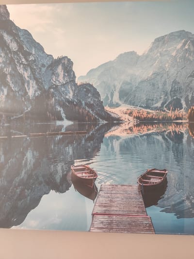 The image depicts a serene mountain lake scene, likely a landscape photograph printed on canvas. The foreground features a weathered wooden dock extending into the calm water. Two small, rustic wooden boats are tied to the dock, one on either side. In the middle ground, a line of similar boats is visible further out on the lake, all appearing empty and at rest.
The background is dominated by imposing, rugged mountains with sharp peaks, some of which are dusted with snow. A band of trees, displaying autumnal colors of orange and brown, is nestled at the base of the mountains on the far side of the lake. The still water perfectly reflects the mountains and trees, creating a symmetrical and peaceful panorama. The sky above is a soft, pale hue, suggesting either early morning or late afternoon, with a gentle, diffused light bathing the entire scene. The overall mood is tranquil and picturesque, evoking a sense of quiet solitude and natural beauty. No people are visible in the image, and there is no visible text.