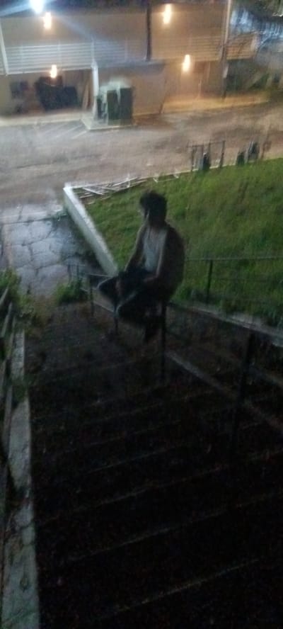 The image shows a dimly lit outdoor scene at night, with a person sitting on a railing. The person appears to be male, wearing a tank top and dark shorts. He is sitting on a dark metal railing that borders a patch of grass. Behind the railing is a grassy slope, and in the background is a building with lights. The stairs in the foreground are dark and lead down out of the frame. The location appears to be in Fort Smith, United States of America.