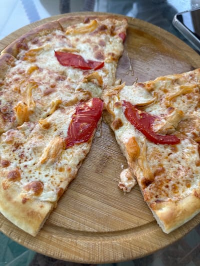 A partially eaten pizza rests on a round wooden board. The pizza is topped with melted cheese, sliced tomatoes, and pieces of chicken. Several slices have been removed, leaving gaps. The wooden board is placed on a glass table. A smartphone is visible in the upper right corner of the image. The scene appears to be an outdoor setting, likely a restaurant, given the presence of a table and a meal. The location is Özgön, Kyrgyzstan.