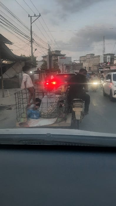 A street scene is depicted under overcast conditions, consistent with dusk or early evening, in RawangItiek, Indonesia. The perspective is from the interior of a stationary vehicle, looking forward through its windshield.

In the mid-ground, a male operator, dressed in a dark shirt and dark pants, drives a motorcycle. A custom-built, open-topped metal cage sidecar is affixed to the motorcycle's left side. Inside the sidecar, an adult individual is seated, partially obscured by light-colored fabric. A young child, clad in a light-colored shirt and shorts, stands unsupported on the sidecar's front left edge, facing forward. Blue bags and miscellaneous items are visible inside the sidecar.

Immediately behind the motorcycle, a dark-colored vehicle displays illuminated red taillights. Additional traffic includes white sedan-type vehicles and another motorcycle with an illuminated headlight, positioned further down the road to the right. The road surface appears to be asphalt.

The background features an array of multi-story commercial and residential buildings, predominantly light-colored concrete constructions. Numerous overhead electrical or communication wires are suspended from a prominent utility pole in the upper left quadrant. A distant communication tower or antenna is visible on the right horizon.