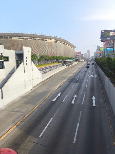 The image depicts a multi-lane avenue in La Victoria, Peru, facing away from the camera. The roadway surface is asphalt with white directional arrows painted on it, along with the word "SALIDA". Vehicles are present on the road. To the left is a large stadium structure, possibly under construction or renovation, with grey walls. Adjacent to the stadium is a modern white concrete structure with stairs. On the right of the avenue, there is a concrete barrier and a series of advertising billboards. In the background, buildings and structures are visible, indicating an urban setting. The sky is clear and pale blue. The scene appears to be taken from a bridge or elevated position, offering an expansive view of the avenue and surrounding cityscape.