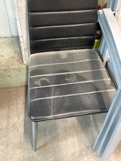 The image shows a dusty, black, padded chair with a metal frame in what appears to be a workshop or storage area. The chair's seat has horizontal indentations and is covered in a layer of dust, with some circular smudges visible. The chair's legs are thin and gray metal. To the right of the chair, there is a portion of a light blue metal structure, possibly another piece of furniture or shelving. In the background, to the left, is a concrete floor and a wall with a textured surface. The overall impression is one of an unkempt, utilitarian space. No people are visible in the image. The specific location in Bad Säckingen, Germany, cannot be determined from the visual information provided.