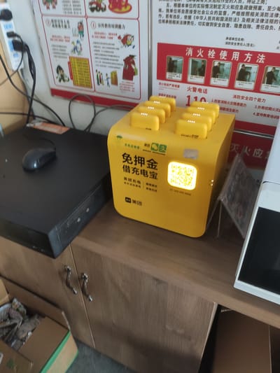 The image depicts an indoor scene, likely within an office or public building in Jianyang City, China. In the foreground, a bright yellow charging station for mobile phones is prominently displayed. This station features multiple charging ports, each with a small, white indicator light, and a large, illuminated QR code on the front for payment or identification. The charging station is branded with Chinese characters, including "免押金充电宝" (deposit-free charging treasure) and "美团" (Meituan, a Chinese e-commerce platform).

To the left of the charging station is a black computer tower with a black mouse resting on top. Cables are visible, including a white power strip with black cords plugged into it, suggesting an active power source. Behind the charging station, on the wall, are several posters with Chinese text and illustrations. One poster, partially visible, contains information related to fire safety, titled "消防栓使用方法" (Fire Hydrant Usage Method) and displays a series of images demonstrating its operation. Another poster on the far left also contains informational graphics and text.

The background shows a wooden cabinet with handles, and a cardboard box filled with various items, possibly discarded materials or supplies, is visible on the floor to the left. The lighting in the scene is consistent with indoor office lighting, and there is no indication of any people actively using the charging station or interacting with the objects in the frame. The overall environment appears organized and functional, typical of a shared space or office setting.