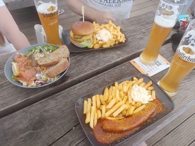 This is a close-up, eye-level, outdoor shot of a wooden table laden with food and drinks. The scene is likely a cafe or beer garden, with a rustic wooden table surface. The lighting suggests daytime, possibly late afternoon.
The table holds two plates of food and three glasses of beer. One plate contains a burger with a toothpick, fries, and mayonnaise. The second plate features a large sausage, covered in a reddish sauce and sprinkled with curry powder, alongside a generous portion of fries. The third dish is a bowl of salmon salad with bread.
Two glasses of Schöfferhofer Weizen beer are prominently displayed. The glasses are tall and slim, filled with golden beer topped with foamy heads. One glass has a label with "Schöfferhofer WEIZEN" and "Saftig-fruchtig!" visible. Another glass, partially out of focus, also shows the "Schöfferhofer" logo. A portion of a third beer glass is visible on the right.
In the background, parts of two people are visible. One person, on the left, has fair skin and is wearing a white top. Another person, in the center-top, is wearing a white t-shirt with text that appears to include "LUCE DEL SUD."
Visible text includes "Schöfferhofer WEIZEN," "Saftig-fruchtig!," "LFORDER QR-Code scannen," "Bestellung aufgeben," "CE 0,51," and "425-0119." The overall impression is a relaxed, enjoyable meal outdoors.