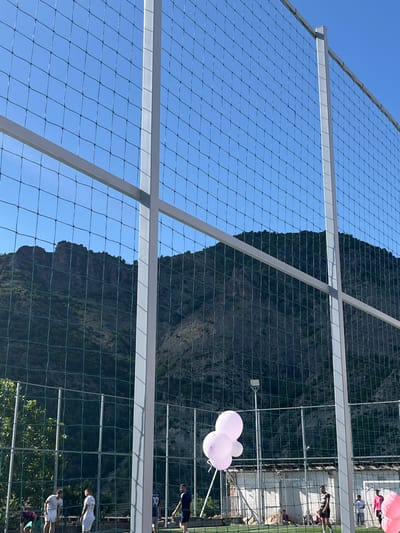 The image shows a mountain backdrop viewed through a net. A white metal frame supports the net. The sky is blue. Pink and white balloons float near the net. People stand near a wall on the right side of the image. The location is Bukovo, Bulgaria.