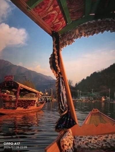 A boat on a body of water with mountains in the background. The boat is ornately decorated with patterned fabric on the ceiling and sides. A traditional Kashmiri boat is docked to the left of the frame. The water is calm and reflects the sky and surrounding scenery. The sky is a clear blue with a few white clouds. The mountains are covered in trees and appear to be a bit hazy. There are a few buildings and other boats visible in the distance on the right side of the frame. The image appears to be taken at sunset, as the sky is illuminated with warm colors.