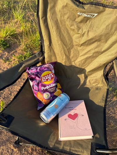 In Sapunov Bor, Russia, a close-up, top-down view shows a foldable camp chair with a khaki-colored fabric seat. Resting on the seat are a bag of Lay's potato chips with a purple and yellow design featuring a salmon image, a light blue can with Russian text, and a pink book with a minimalist drawing of a person and a heart. The chair is positioned on dry, sandy ground with sparse green grass and vegetation visible in the background. Sunlight casts shadows on the chair's fabric, suggesting it is daytime. The overall scene depicts a relaxed outdoor setting.