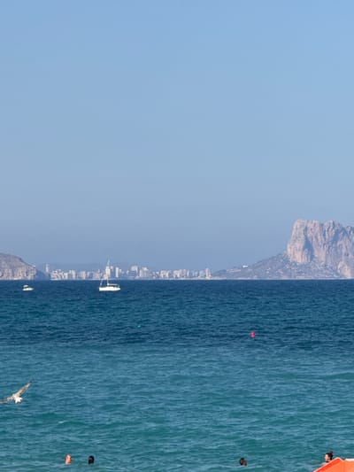 This is a landscape photograph of the coast near Pla Parcial Platja, Spain, under a clear, pale blue sky. The image is divided into three horizontal sections: the sky, the sea, and a distant cityscape with a prominent rock formation.

In the foreground, the deep blue sea is in motion with moderate waves and ripples. A seagull is captured in flight, its wings extended, over the water. Below the seagull, two individuals are partially submerged in the sea, with only their heads visible. To the right, another person is visible in the water near an orange object, possibly a life raft or a buoy. A red buoy is also visible further out in the water.

The middle ground features the vast expanse of the Mediterranean Sea, with two sailboats navigating the water. One is a small white boat with a single mast, and the other is a larger white sailboat with its sail raised. In the distance, a city skyline stretches along the coast, characterized by numerous buildings. To the far right of the cityscape, a large, rugged rock formation dominates the horizon, jutting out into the sea. The overall scene depicts a serene coastal environment with recreational activities and a distinctive geographical landmark.