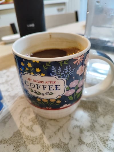 A ceramic mug, dark blue with a floral pattern of pink, yellow, and light blue flowers and foliage, stands prominently on a light-colored, textured tablecloth. The mug displays a white banner containing the red text "LIFE BEGINS AFTER" above the blue text "COFFEE". The bottom segment of the mug is white with a thin red line, and it features a white handle on the right side. Inside the mug is a dark brown liquid consistent with coffee, exhibiting a light brown crema or foam layer on its surface. In the blurred background, a tall, cylindrical black electronic device, possibly a speaker with an "LG" logo, is visible to the left. To the right, a clear glass vessel, possibly a carafe or pitcher, with condensation droplets on its exterior, is present. The scene is observed indoors, likely a domestic kitchen or dining area, within the Municipal Unit of Acharnes, Greece.