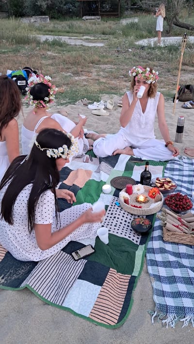 Four women are sitting on a blanket on the sand near Agia Triada, Greece. Three of the women are wearing flower crowns. The woman in the center is drinking from a white cup. There are various items on the blanket, including a bottle of wine, a bowl of cherries, a bowl of sliced fruit, a woven basket, and some rolled-up items. A cell phone is lying on the blanket. In the background, there is some dry grass and a partially visible structure. One woman is standing in the distance.