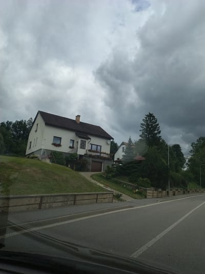 The image shows a white, two-story house situated on a sloped grassy hill. The house has a dark-colored roof and a brick chimney. It features several windows, some with flower boxes. A driveway leads to a garage door on the lower level of the house. A path curves from the driveway up to the house. In the foreground, a paved road runs alongside the property, with a concrete retaining wall separating the road from the grassy slope. The sky above is overcast with dark, heavy clouds. The scene appears to be a residential area in Olešná, Czechia, given the context. There are no people visible in the image.