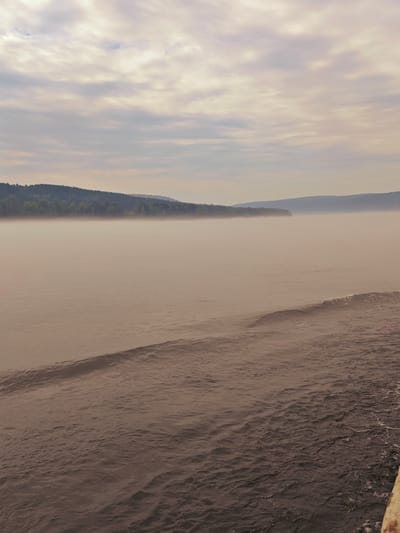 The image depicts a tranquil, misty river scene in Atamanovo, Russia. The foreground is dominated by the rippling surface of the water, showing subtle waves and a wake. In the midground, a thick layer of fog partially obscures the distant shoreline, which is lined with a dense, dark green forest. The background features rolling hills fading into the hazy distance under a cloudy sky. The overall atmosphere is serene and slightly melancholic, with muted colors and soft lighting contributing to the impression of an early morning or late evening. There are no people or man-made structures visible in the scene, emphasizing the natural beauty of the location.