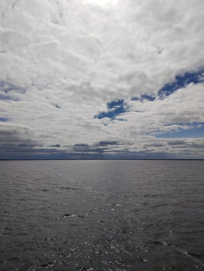 The image presents a wide, expansive view of a large body of water under a dynamic sky, located in Pochinok, Russia. No people or specific objects are visible, emphasizing the natural landscape.
The upper half of the frame is dominated by a sky filled with layered, mostly white and light grey clouds. These clouds appear to be stratocumulus or altocumulus formations, with some brighter areas suggesting the presence of sunlight behind or above them. Through breaks in the cloud cover, particularly towards the right and center-left, patches of clear blue sky are visible.
A distinct, flat horizon line cleanly separates the cloudy sky from the water below. There's a very faint, dark line just above the horizon on the water side, which could indicate a distant landmass or shoreline.
The lower half of the image features a vast expanse of dark grey water, likely a lake or a wide river. The water's surface is not entirely smooth, displaying numerous small ripples and gentle waves. These ripples catch and reflect the ambient light, creating a shimmering, textured effect across the otherwise dark water. The scene conveys a sense of serene vastness and natural grandeur, captured during the daytime under variable cloud conditions. There is no visible text in the image.
