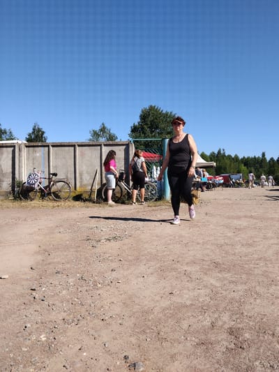 A medium shot captures an outdoor scene with a clear blue sky and a dusty, gravel-strewn ground. In the foreground, a woman wearing a black tank top, black leggings, and pink and white sneakers walks towards the camera. She has short brown hair and is wearing a red visor. A black fanny pack is around her waist. Behind her, to the left, is a concrete wall. Two bicycles are leaning against the wall. One bicycle on the far left has a dartboard attached to its frame. Two women are standing near the bicycles, one wearing a pink t-shirt and denim shorts, the other wearing a black and white striped shirt and black shorts. Further in the background, there are several market stalls with tents and vehicles, and a few more people are visible. The scene appears to be a market or an outdoor event.