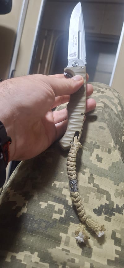 A person's hand holds a tan-handled folding knife. The blade is partially open and made of polished metal. Engraved text on the blade appears to read "KTTAP" and "HOK". A braided tan cord is attached to the knife's pommel and hangs down, featuring a decorative metal bead with a silver Celtic-like design and a golden element. The person is wearing camouflage pants. The background is out of focus but suggests an indoor setting with vertical elements. The location is identified as Orlivschyna, Ukraine.