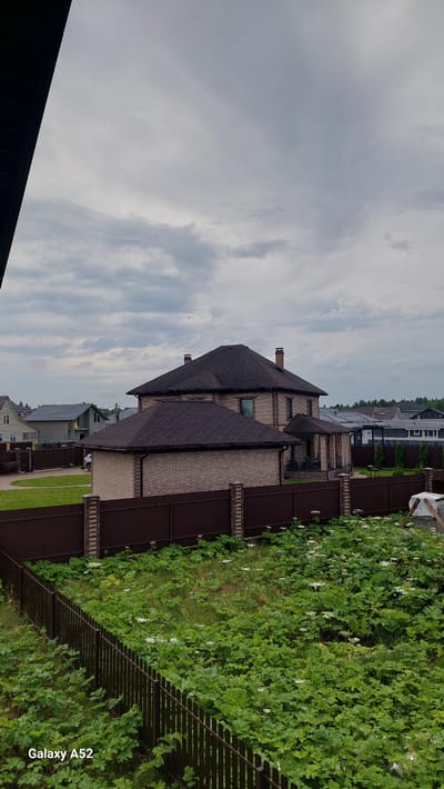The image captures a suburban scene in the city of Dolce Vita KP, Russia, on a cloudy day. The foreground is dominated by a dark, slatted fence and an overgrown patch of green vegetation with small white flowers, hinting at a somewhat unkempt garden or natural area. Beyond the fence, a brown brick house with a dark, shingled roof is visible, featuring multiple windows and a chimney. To the left of this main house, a smaller, attached brick structure with a similar roof is present. Further in the background, other houses with diverse architectural styles and roofing materials can be seen, along with more fencing and greenery. The sky is overcast with diffused light. A car is partially visible in the driveway of one of the properties. The "Galaxy A52" watermark is present in the bottom left corner, indicating the device used for capture.