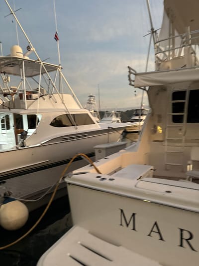 The image shows the stern of two white boats docked side-by-side in a marina located in Playa Herradura, Costa Rica, at coordinates 9.651 latitude and -84.665 longitude. The boat on the right has the word "MAR" in black letters on its stern. Both boats have a yellow hose attached to their sides. The boat on the left has a white radar dome on its top. There are several other boats in the background. The sky is blue and cloudy. The water is dark and there is a buoy in the foreground. The scene appears to be taken at dusk.