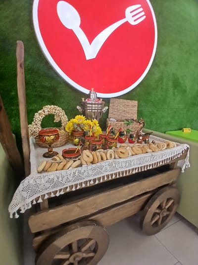 The image depicts a wooden cart laden with a traditional Russian spread, possibly for a celebration or event. The cart is positioned in front of a green, textured background, reminiscent of artificial grass. Above the cart, a large, circular red sign with white outlines of a spoon and a fork is mounted.

The cart itself is made of rustic wood, with visible grain and a weathered appearance. It has two large, spoked wooden wheels on the right side, and the left side is partially obscured by a wooden pole. The top of the cart is covered with a white, lacy tablecloth that hangs over the edges, adorned with decorative tassels.

Arranged on the cart are several items characteristic of Russian hospitality. In the center, a silver samovar stands prominently, a traditional device for heating water for tea. Beside it, a woven basket contains a ring of small, ring-shaped bread rolls known as "baranki" or "sushki." A decorative basket with painted motifs holds more of these rolls. There are also several small, painted jars, likely for jam or honey, and a tall, stemmed glass with intricate red and gold painting. Further to the right, a group of traditional Russian nesting dolls (Matryoshka dolls) are arranged, along with other small decorative figures, including what appears to be a swan. The main focus of the cart's display is a generous assortment of baranki and other similar baked goods, arranged in a pleasing manner. Yellow sunflowers add a touch of color to the arrangement.

The lighting is even, suggesting an indoor setting. There are no people visible in the image. The overall impression is one of a carefully curated display of traditional Russian food and cultural items. The presence of the food items and the cart suggest a festive or welcoming atmosphere. The location in Knyazevo, Russia, would be consistent with such a display of national culinary traditions.