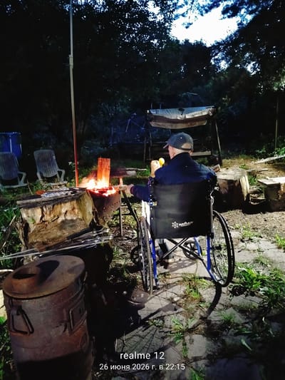 A person in a wheelchair is sitting in an outdoor setting at night, facing a bonfire. The person is wearing a dark jacket and a baseball cap. They are holding a yellow object in their right hand. The wheelchair has "АРМЕД" printed on the back. In the foreground, there is a large, rusty metal stove. A bonfire is burning in a metal fire pit, with logs and skewers nearby. Two empty lawn chairs are visible in the background, along with trees and foliage. The ground appears to be a mix of grass and a paved or concrete area. The timestamp in the lower right corner indicates "realme 12 26 июня 2026 г. в 22:15", which translates to "realme 12 June 26, 2026 at 22:15". The location is identified as Novoe, Russia.
