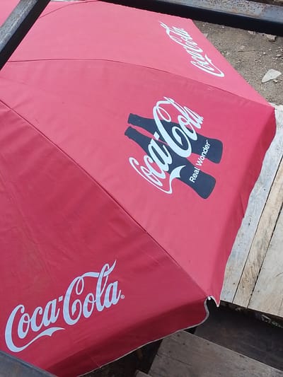 The image shows a red Coca-Cola branded umbrella, likely on display at an outdoor vendor stall in Abeere, Nigeria. The umbrella is opened and angled, revealing the iconic white Coca-Cola logo printed multiple times in a script font. Two stylized black bottles are also depicted on the umbrella, with the text "Real Wonder" written between them. The background consists of a rough, stony ground, and some wooden planks that appear to be part of the vendor's setup. A dark metal bar runs along the top edge of the image, possibly part of the umbrella's frame or a nearby structure. The scene suggests a commercial setting where Coca-Cola products are sold.