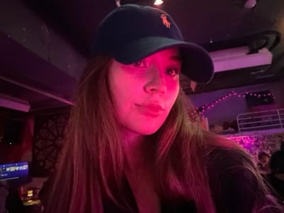 The image depicts an indoor scene in 마포구, South Korea, characterized by dim lighting and prominent pink illumination. A person, identified by long, light brown hair and a dark blue baseball cap with a light-colored polo horse logo, occupies the foreground, facing the camera. Their face is partially illuminated by a strong pink light source, highlighting their complexion and features. In the background, the environment is sparsely lit with purple and pink ambient lights. Visible elements include a decorative circular pattern on a wall to the left, a television screen or monitor further left, and a white railing structure in the mid-background. String lights are visible in the upper background, and several indistinct figures of other individuals are present in the deeper background. The overall setting suggests a bar or club.