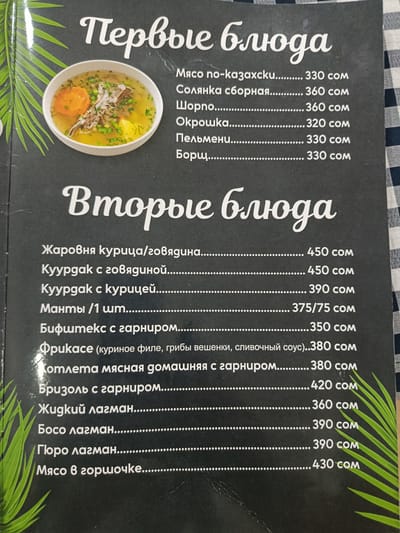 The image displays a menu board with a list of dishes and their prices. The menu is divided into two sections: "Первые блюда" (First Courses) and "Вторые блюда" (Second Courses). Under "First Courses," items include "Мясо по-казахски" (Kazakh-style meat), "Солянка сборная" (Assorted Solyanka), "Шорпо" (Shurpa), "Окрошка" (Okroshka), "Пельмени" (Pelmeni), and "Борщ" (Borscht), with prices ranging from 330 to 360 som. The "Second Courses" section lists items such as "Жаровня курица/говядина" (Roaster chicken/beef), "Куурдак с говядиной" (Kuurdak with beef), "Куурдак с курицей" (Kuurdak with chicken), "Манты /1 шт" (Manti /1 pc), "Бифштекc с гарниром" (Beefsteak with garnish), "Фрикасе (куриное филе, грибы вешенки, сливочный соус)" (Fricassee (chicken fillet, oyster mushrooms, cream sauce)), "Отлета мясная домашняя с гарниром" (Homemade meat cutlet with garnish), "Бризоль с гарниром" (Brizole with garnish), "Жидкий лагман" (Liquid lagman), "Босо лагман" (Boso lagman), "Гюро лагман" (Guro lagman), and "Мясо в горшочке" (Meat in a pot), with prices ranging from 350 to 450 som. There is a picture of a bowl of soup on the left side of the menu. The background is a dark, textured surface. The menu board appears to be in a restaurant setting. The location in the city of Kara-Balta, Kyrgyzstan, is not visually indicated by any landmarks or signs within the image.