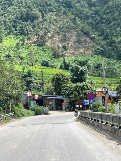 The image depicts a roadside scene in Binh Lu, Vietnam, dominated by a lush, green, terraced hillside and dense forestation. A paved road curves through the foreground, bordered by a metal railing on the right. On the left side of the road, a small cluster of buildings, likely shops or residences, are visible, adorned with Vietnamese flags. One building has an open storefront displaying goods. A motorcyclist is seen riding away from the viewer on the road. Traffic signs, including a blue directional sign and a red circular prohibition sign, are present near the buildings. The environment appears rural, with evidence of agriculture on the hillside.