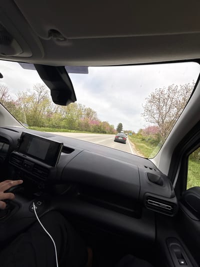 The image shows the interior of a vehicle, likely a car, in motion on a road. The view through the windshield reveals a two-lane road with a black car ahead. The surroundings are characterized by trees and green foliage with hints of pink blossoms, suggesting a springtime setting. Inside the vehicle, the dashboard is visible, featuring a screen and ventilation controls. A hand is seen near the controls, and a charging cable is plugged into a port. It is not possible to determine the precise location within Varbak, Bulgaria.