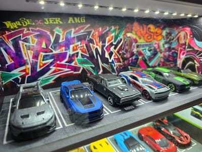 The image displays a collection of toy cars arranged on shelves, with a graffiti-style backdrop. The graffiti features bold, colorful lettering and abstract designs. The toy cars are detailed models, some appearing to be muscle cars and sports cars, and are parked in designated spots on the shelves. The scene is illuminated by a strip of LED lights located above the graffiti backdrop. There are no people visible in the image. The specific location of Puhoi, Moldova, is not discernible from the visual content of the image.