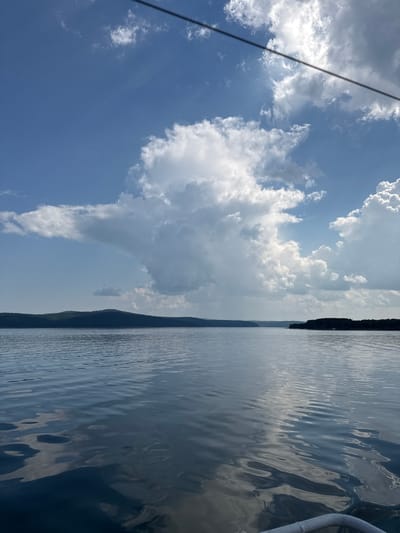 This is a vertical, full-shot photograph taken from a boat on a body of water, likely a lake or large river, under a bright, partly cloudy sky. The water is calm with gentle ripples reflecting the sky and clouds. In the distance, a dark, tree-covered shoreline forms a horizontal band across the image. The sky is a vibrant blue, dotted with large, fluffy white clouds, some of which are illuminated by sunlight. A thin, dark wire stretches diagonally across the upper portion of the frame. The scene appears to be natural and tranquil.