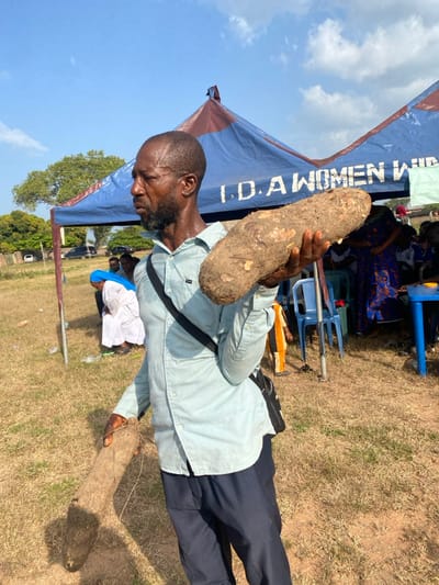 An outdoor scene in Itobe, Nigeria, depicts a dark-skinned adult male of apparent middle age standing centrally, holding two large root vegetables. The man wears a light blue long-sleeved collared shirt, dark blue trousers, and a black shoulder bag. His facial expression is neutral, and his gaze is directed to his left. He holds a large, brown, oblong yam horizontally in his right hand and a longer, thinner, brownish root vertically in his left hand, extending towards the ground.
The environment consists of a dry, yellowish-brown grassy field with patches of exposed earth under a clear blue sky with sparse white clouds. In the background, two canopy tents are visible, one predominantly blue with a red/maroon trim and white lettering that reads "I.D. A WOMEN WING". Under and around the tents, several individuals are present, including one person in a white robe and blue head covering seated on the ground to the left, and others partially visible sitting on blue plastic chairs or standing. A blue plastic table is partially visible to the far right. Distant green trees and parked vehicles are discernible on the left side of the background.