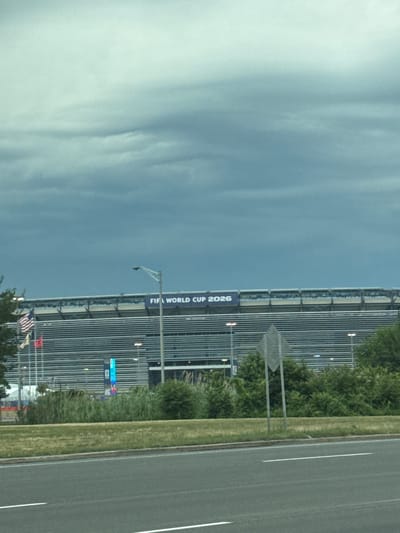 The image shows a stadium in East Rutherford, New Jersey, United States of America, with a banner that reads "FIFA WORLD CUP 2026". The stadium is large and modern, with rows of seating visible. There are American flags and other flags flying in front of the stadium. The sky is cloudy, and the overall lighting suggests an overcast day. A road and some greenery are in the foreground.