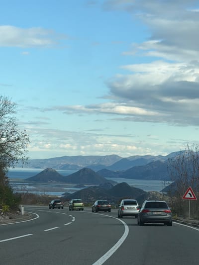 The image depicts a winding road in Donji Ulići, Montenegro, with a line of cars traveling along it. The road is bordered by a view of mountains and water, likely a lake or the sea. A road sign indicating a sharp left curve is visible near the front of the car line. The sky is partly cloudy, suggesting fair weather.