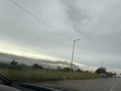 This is a photograph taken from inside a vehicle, looking out through the windshield, which has visible water droplets and streaks, suggesting it is raining or has recently rained. The scene depicts a rural or semi-rural setting under a cloudy, overcast sky.
In the foreground, the wipers of the vehicle are visible at the bottom of the frame. The middle ground features a stretch of grassy vegetation, with scattered trees. Two streetlights on poles are visible along a road that appears to be paved with cobblestones or rough material.
Further in the background, there are two cars on the road. The car on the left is a dark sedan, and the car on the right is a lighter-colored vehicle, with its headlights on. Beyond the road and vegetation, a distinct, dark, smoky band stretches across the horizon, partially obscuring the sky. This band could indicate smoke from a fire or industrial activity. The sky is predominantly gray and cloudy, with some lighter patches suggesting the sun is trying to break through or that there is a break in the cloud cover in the distance.
The overall mood of the photograph is somewhat somber due to the weather conditions and the smoky haze. The time of day appears to be either early morning or late afternoon, given the dim lighting. The location context of Ivanyane, Bulgaria, suggests this is a landscape in that region. No people are clearly visible in the photograph.