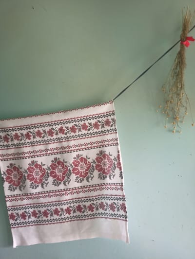 A close-up, eye-level shot shows a white embroidered fabric hanging against a pale green wall. The fabric, which appears to be a decorative towel or textile, features a repeating pattern of red and gray floral motifs and geometric borders. The embroidery is intricate, with small stitches forming the designs. The fabric is draped and secured by a dark string that runs diagonally across the wall. To the right of the fabric, a small bunch of dried, wispy plants with tiny yellow flowers is tied with a red ribbon and hangs from the same string. The wall itself is textured and shows some minor imperfections and dust. The scene is lit with soft, natural light, creating subtle shadows. This image depicts a rustic interior setting.