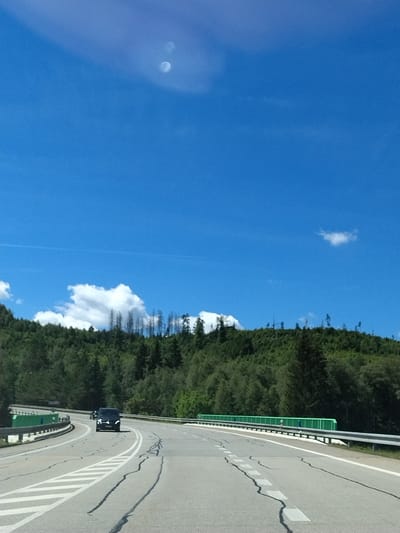 The image depicts a highway curving through a forested mountainous landscape, likely in the vicinity of Strážný, Czechia, given the provided context. The sky is a bright, clear blue with a few wispy clouds and two bright circular artifacts, possibly lens flare or reflections. A dense green forest covers the hillsides, with some areas showing patches of dead, bare trees, suggesting past deforestation or natural events. The highway itself is paved with visible cracks and markings. On the road, a dark-colored SUV is driving away from the viewer, and a motorcycle is visible behind it. Guardrails line the sides of the highway, with a distinctive green barrier visible on the right side of the road. The overall scene suggests a sunny day on a mountain road.