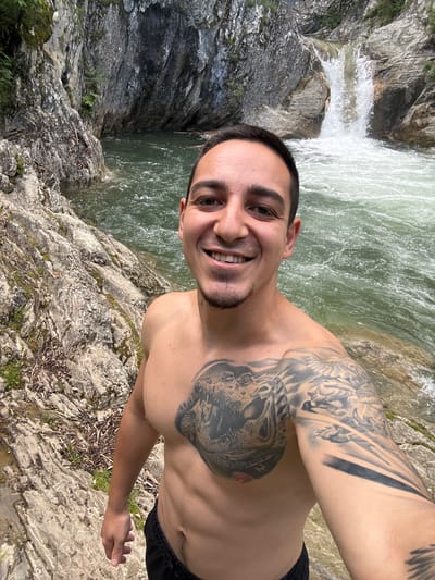 A shirtless man with dark hair and a tattoo on his chest and arm is standing on a rocky surface near a waterfall. The location is in Medven, Bulgaria. The man is smiling and appears to be taking a selfie. The background includes a waterfall, rocks, and vegetation. The water is a green-tinted white due to the falls.