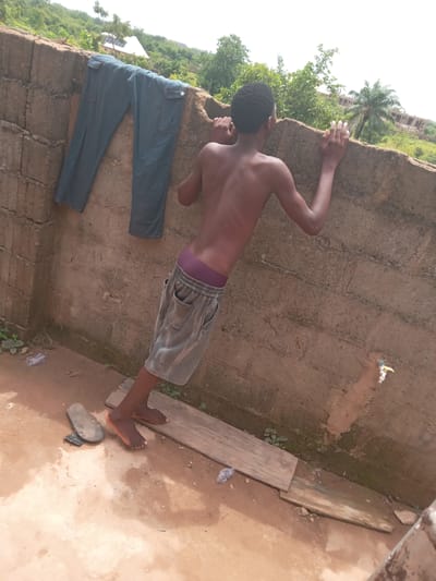 The image depicts an individual standing barefoot in an outdoor setting in Achara Ndiagu Omegio, Nigeria. The person, a young male, is shirtless and wearing shorts with a visible elastic waistband. He is leaning against a weathered, uneven wall, possibly constructed of bricks or concrete. A pair of blue jeans is draped over the wall to the left of the individual. Beside his feet is a worn-out, broken sandal. The ground appears to be dirt or a similar natural surface. Wooden planks are present on the ground near the wall. Vegetation and trees are visible in the background, suggesting a rural or semi-rural location. A water tap is affixed to the wall.