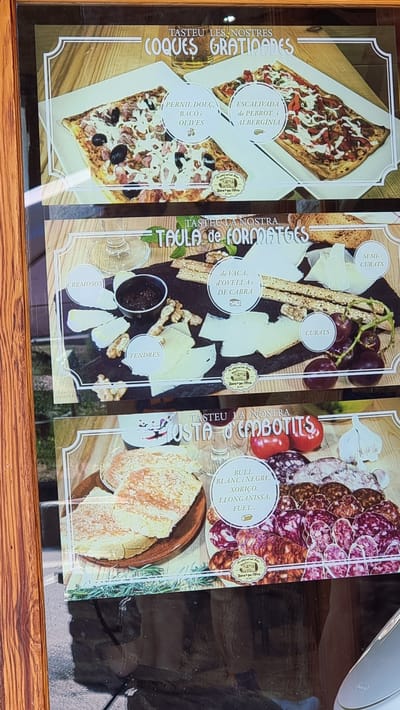 The image is a photograph of a menu display board for a restaurant in Rupit, Spain. The display features three sections showcasing different food items.
The top section, titled "TASTEU LES NOSTRES COQUES GRATINADES," displays two types of gratinated flatbreads. The one on the left is topped with ham, bacon, and olives. The one on the right appears to be an escalivada, with roasted red peppers and eggplant.
The middle section, titled "TAULA de FORMATGES," shows a cheese platter. Various types of cheese are arranged on a dark surface, accompanied by crackers, walnuts, grapes, and a small bowl of jam. The cheeses are labeled as "CREMOSOS" (creamy), "TENDRES" (tender), "DE VACA, D'OVELLA i DE CABRA" (from cow, sheep, and goat), "SEMI-CURATS" (semi-cured), and "CURATS" (cured).
The bottom section, titled "TASTA D'EMBOTITS," presents a selection of cured meats. The display includes bread, tomatoes, garlic, and an assortment of sliced sausages. The sausages are described as "BULL BLANC i NEGRE, XORIÇO, LLONGANISSA, FUET..." (white and black sausage, chorizo, longanissa, fuet).
Each section includes a small circular emblem with text, likely indicating the restaurant's name and location. The overall presentation suggests a rustic and traditional culinary offering.