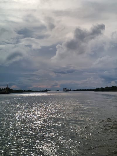 This is a photograph taken from a body of water, likely a river or bay, looking towards a distant shoreline. The sky is overcast with dramatic, heavy clouds, but there are patches of sunlight breaking through, casting a shimmering effect on the water's surface. The water itself is dark and slightly choppy, with reflections of the sky and sunlight scattered across it.
On the left side of the image, near the shore, there is a large crane, suggesting a port or industrial area. Further along the shoreline, there are trees and buildings visible. In the distance, several boats are present on the water. One larger vessel, a white passenger ship with multiple decks, is prominently visible in the middle ground, moving away from the viewer. Another, smaller boat is seen to its left. The shoreline stretches across the horizon, with greenery and some structures dotting the landscape. The overall atmosphere is somewhat moody due to the clouds, but the sunlight on the water adds a touch of brightness and dynamism to the scene.
This scene is located in Kargulino, Russia.