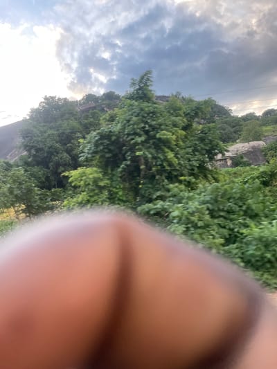 The image, taken in Igbo Ijaiye, Nigeria, shows a blurry foreground of what appears to be skin, possibly a hand or arm, obscuring the view. Beyond this foreground, a lush, green landscape unfolds, with dense vegetation and trees covering a hillside. In the background, a rocky outcrop is visible, and the sky is filled with dramatic, cloudy formations, suggesting an overcast or stormy weather condition. The overall impression is of a natural, outdoor setting, partially obscured by the photographic subject.