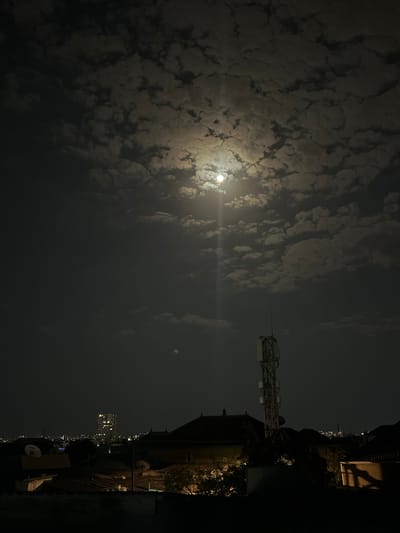 The image depicts a nighttime scene in Sabunchu, Azerbaijan, dominated by a full moon partially obscured by scattered clouds. The moon casts a bright glow through the clouds, creating a luminous effect in the dark sky. In the foreground, silhouetted rooftops of various buildings are visible, with some satellite dishes present. A tall telecommunications tower stands prominently on the right side of the frame, with lights illuminating its structure. In the distance, a modern high-rise building with illuminated windows is visible against the city skyline, suggesting an urban environment. The overall atmosphere is serene and atmospheric, with the moonlight being the primary light source. No people or actions are discernible in the image.
