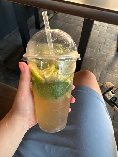 An overhead shot captures a person's hand holding a clear plastic cup filled with a bubbly, light brown beverage. The drink contains slices of lime and green mint leaves, suggesting a refreshing cocktail or mocktail. The cup has a domed lid with a plastic straw inserted. The person is wearing blue shorts. In the background, a dark wooden tabletop is visible, along with a textured tiled floor. Part of a stroller is also in view on the right side of the frame. The scene appears to be outdoors or in a café with outdoor seating. This image was taken in Sarny, Ukraine.