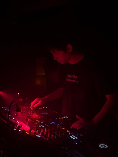 The image displays a male individual, positioned in profile from the torso upwards, actively operating professional DJ equipment within a dimly illuminated, predominantly red-lit environment. The subject is wearing a dark t-shirt inscribed with the text "VACUUM 1st Anniversary" in red lettering. Both hands are engaged with the multi-deck DJ console, manipulating various controls including jog wheels, faders, and a multitude of illuminated buttons emitting red, orange, green, and blue light. A gooseneck lamp is positioned over the mixer, providing focused light. The background is largely indistinct and dark, characteristic of a nightclub or music venue. The specific location within the Changning District, China, is not discernible from the visual evidence provided.