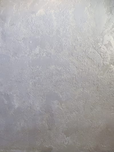 The image is a close-up, vertical, full-frame shot of a textured wall with a light gray or silver finish. The texture is uneven, with raised and recessed areas that create a stucco-like or Venetian plaster appearance. The lighting creates subtle highlights and shadows, emphasizing the depth and variation of the surface. There are no people, objects, or discernible environmental features visible. The location is not specified within the image itself.