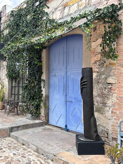 The image shows a building facade in San Miguel de Allende, Mexico. A blue arched double door is visible, with the inscription "Filippo Giusti, Visual Artist" on the door. A dark metal sculpture of a hand holding a cylinder is mounted on a black pedestal beside the door. The building exterior is constructed of stone and brick, with ivy growing on the walls and around a barred window below the door. The image appears to have been rotated 90 degrees.