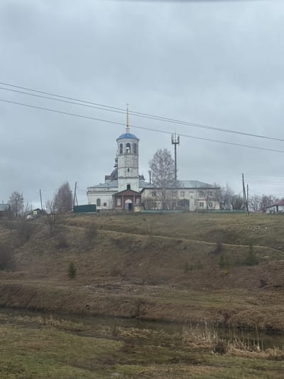 The image depicts an overcast, daytime scene in a semi-rural setting, likely in late autumn or early spring given the bare trees and dry vegetation. A prominent white church building, characteristic of Eastern Orthodox architecture, stands in the mid-ground. It features a multi-tiered bell tower topped with a blue, onion-shaped dome and a golden cross. The church appears weathered, suggesting age.
To the right of the church, a steep hill rises, covered in dry, brown, and some greener grass, indicating a dormant or sparse landscape. Scattered residential buildings, some appearing aged, are visible behind and around the church, nestled among bare deciduous trees.
Overhead, utility poles and multiple power lines crisscross the gray sky. A modern cellular tower with antennae is also visible in the background, contrasting with the older architecture of the church. On the ground, near the base of the church, a yellow diamond-shaped road sign featuring a pedestrian crossing symbol is clearly visible. No people or specific activities are present, contributing to a quiet, still atmosphere. The uniformly cloudy sky suggests an overcast day. The location is Богородск, Russia.